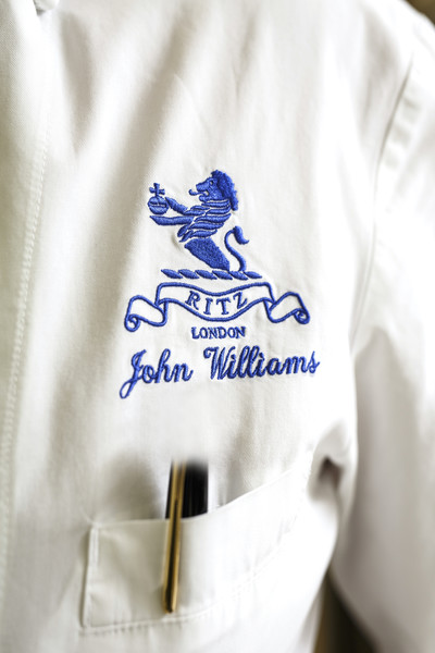 Close-up of a white chef's jacket embroidered with 'Ritz London' and the name 'John Williams' in blue, worn by John Williams, Executive Chef at The Ritz London. A pen is tucked into the pocket beneath the embroidery.